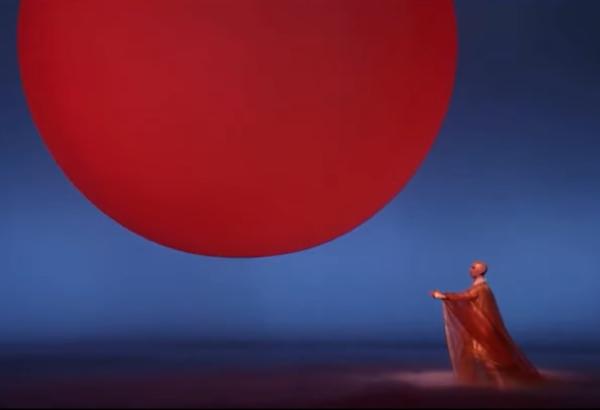 A deep red semi-circle spans most of the frame, cut off at the top of the frame just above its equator. The background is shades of blue, darker at the top.  A bald figure in orange robes and a white collar stands below and to the right of the sun, facing left, golden-color head tilted slightly up, and left arm raised to horizontal, in supplication probably, trailing filmy orange robe extending to the wrist, with a bright hand of uncertain blobby shape.  The figure is Anthony Roth Costanzo on stage in the opera Akhnaten, performing Hymn to the Sun.