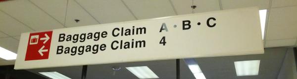 A sign hanging from a drop ceiling with fluorescent lighting fixtures.  There are two rows of text, in a medium-weight Helvetica-like font.  To the left of the text, in a red box with white text, are two rows of symbols, aligned with the two rows of text.  The first row has a baggage icon, and a bold white arrow pointing to the right.  The second row has a bold white arrow pointing to the left.  The first row of text reads, Baggage Claim A · B · C.  The second row reads, Baggage Claim 4.