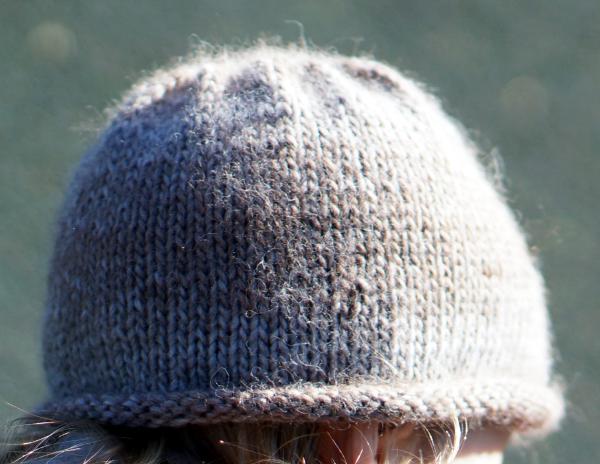 A plain, gray wool hat, worn by is probably a person but only a little strip of head is visible between the bottom of the hat and the bottom of the picture, showing hair and possibly light-colored skin on one side.  The background is plain and blurry, with some bokeh circles in the top corners.