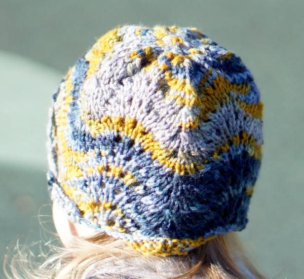 A knitted hat with a complicated pattern reminiscent of the top of the Chrysler Building, with stripes of yellow, light blue, and dark blue color, worn by a child or baby facy away, with the right side in shadow, and light-colored straigh hair wisping sideways below the hat.