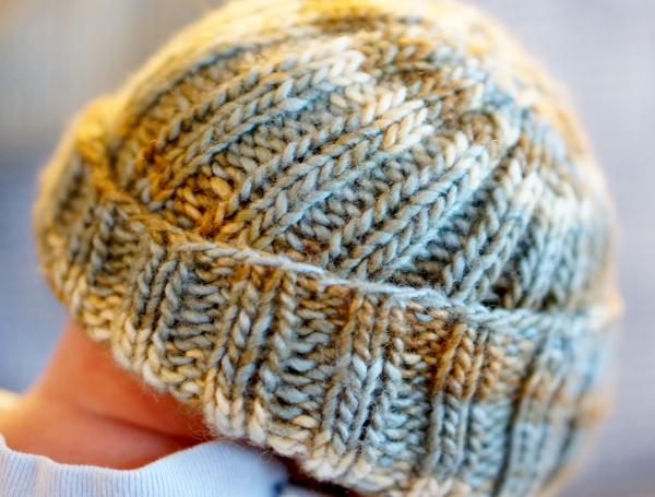 A baby hat, on a baby's head, a little bit of neck visible.  The brim of the hat is folded up crookedly.  The hat is ribbed 2k2p in medium yarn, in swirling cream, light brown, and light blue colors.