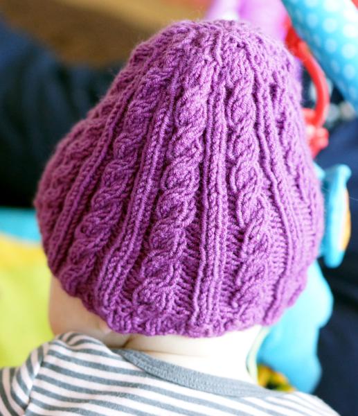 a purple knit hat in alternating ribs and braids, slightly pear-shaped, worn by a baby facing away, a striped white and gray shirt visible, with a background out of focus.