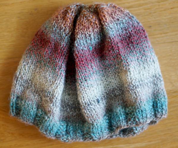 A knit hat lying flat on a wood background, plain-knit with a ribbed edge, in spiral colors of brown, light blue, light scarlet, and white.