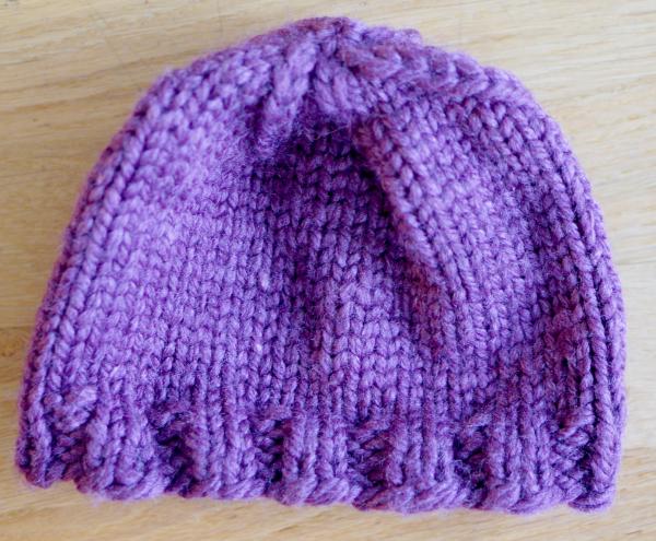 A purple knit hat lying flat on a wood background, plain knit with a ribbed edge.  The yarn appears large, or else the hat is small.