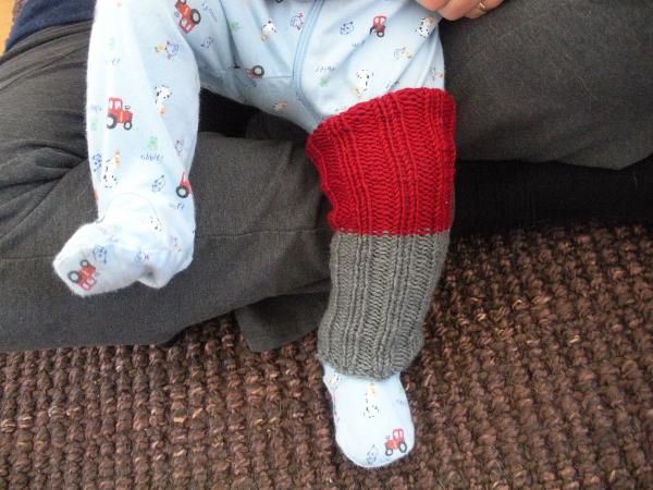 A baby sitting in a larger person's lap, from their waists down.  The larger person is wearing gray sweatpants. The baby is wearing a light blue jumpsuit decorated with little tractors, cows, and other farm images. Their left leg extends to the floor, jumpsuited foot on the coir rug, nicely displaying a knitted gray and red legwarmer over the jumpsuit.