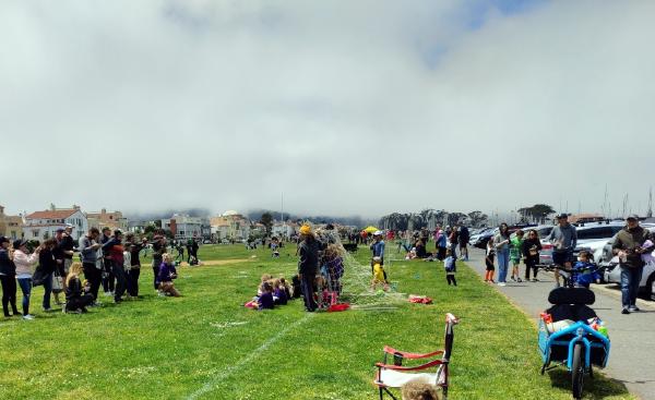 The corner of a sunny, verdant childrens' soccer field in the foreground, with many people in the midground. A fogbank in the distance covers the top half of the picture, with a few tiny bits of blue sky visible.  Some ship masts are visible at the right edge, above parked cars and people walking on a sidewalk.