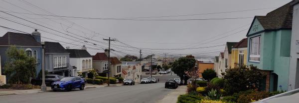 A downhill-sloping residential road, with row houses on both sides and a gray sky in the background, cluttered by dozens of utility wires.