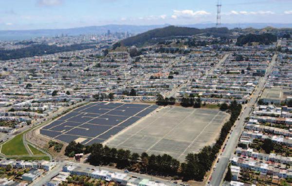 An aerial view of northern San Francisco, pointing east.  A large rectangular area fills the foreground, with one side cement-colored and the other solar panels.  A grid of houses fills the middle third, with a mountain in the top middle and a tower on top of the mountain.