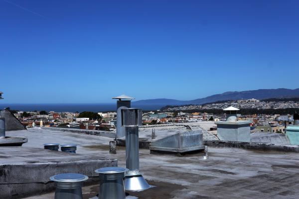 A rooftop fills the bottom third of the rooftop, with a dirty gray surface and a scattering of shiny metal pipe tops and exhausts.  Above the roof, a thin line of houses and patches of tree extends across the picture.  Aobev that, on the left side, a stretch of blue water with a flat horizon.  On the right side, the water disappears behind hills, and on the far side of the water, a ridge line emerges from the water and extends above the nearer hills to the edge of the picture.  The top half of the picture sky shading from light blue up to a medium and rich blue at the top.  The sky is cloudless except for one contrail streaking faintly from the top left corner, fading out before reaching the middle of the picture.  Very dark shadows extend briefly below some of the tops of the roof objects.