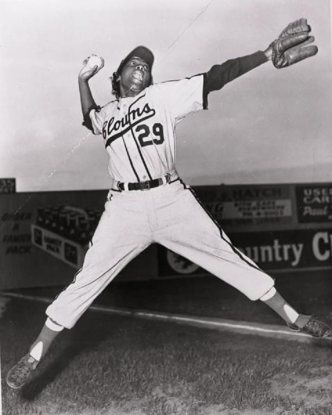 Black and white photograph of a right-handed baseball player in a light-colored unifom with 29 Clowns on the front, jumping with legs split and not touching the ground, holding a baseball in their hand with their arm cocked back, and the other, gloved hand pointing straight forward.  Their mouth is open in a shout.