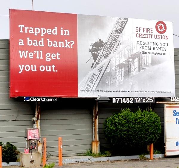 A billboard on the side of a two-story building.  The left side is red, with white text reading, Trapped in a bad bank?  We'll get you out.  The rest of the billboard is a grayscale image of a firefighter climbing a large ladder extending to the top of the billboard, with a spray of water passing left to right below them, and a burning, scaffolded building on the right.  Red text overlays the image in the top right, SF FIRE CREDIT UNION Rescuing you from bad banks. sffirecu.org/rescue. Disclaimer text at the bottom: Privately insured by ASI.  This institution is not federally insured.  The credit union is not insured by any state government.  The bottom edge of the billboard including the logotype Clear Channel and the text #71452 12'x25'.